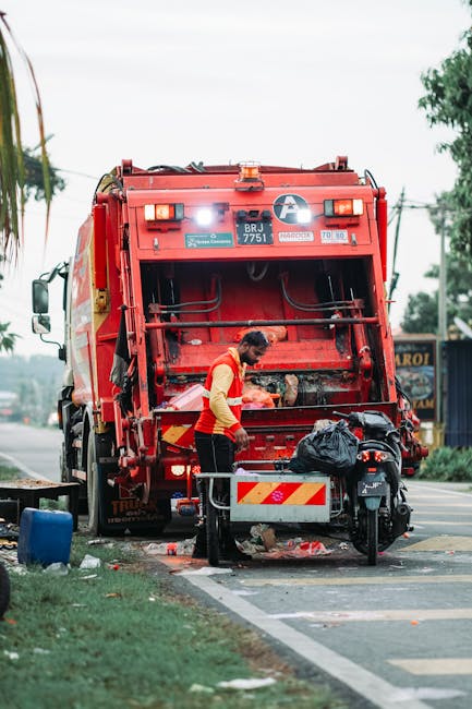 A sanitation worker, dressed in a high-visibility yellow and orange uniform, is seen operating the rear mechanism of a large red rubbish collection truck parked on a street. The truck, equipped with hydraulic lifting equipment, is positioned next to the curb, with its rear open and filled with waste. The worker appears to be in the process of clearing or sorting waste, with a black garbage bag placed on a small metal platform attached to the truck. The surrounding environment includes a paved sidewalk, a grassy strip, and some scattered debris or waste on the ground nearby. In the background, there are trees, utility poles with power lines, and a building with signage partially visible on the right side. The scene is illuminated by natural daylight, creating a clear and observational view of an on-site waste collection activity, which may be connected to private or independent rubbish removal services like Waste Removal Harrow, demonstrating the process of on-site waste handling outside of municipal collection.