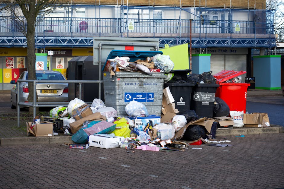 A large collection of overflowing waste bins and surrounding rubbish is situated on a paved area of a commercial or retail parking lot. The central focus is on an open, gray mixed waste bin filled with crumpled papers, cardboard, and various discarded packaging. To its right are black and red recycling bins, with black bin bags and other waste materials piled on and around them, including flattened cardboard boxes, plastic bottles, and paper debris. The area around the bins features scattered rubbish, such as flattened boxes, plastic packaging, and loose paper. In the background, there is a building under renovation or construction, covered with blue scaffolding and safety signage. To the left, a silver car with a partially visible license plate is parked close to the rubbish collection point, separated from the waste by a metal railing. The scene is lit by natural daylight, with minimal shadows, illustrating a typical example of improper waste disposal or insufficient rubbish collection, relevant to private waste removal services provided by Waste Removal Harrow.
