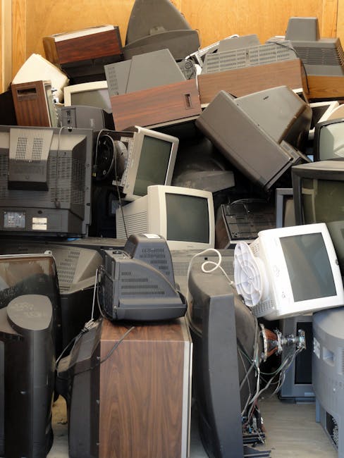 A large collection of old, unused electronic devices, primarily consisting of vintage CRT computer monitors, each with curved glass screens and plastic casings in shades of grey, black, and beige. The monitors are arranged in a somewhat haphazard manner, stacked against a wooden wall and piled on top of each other. Some monitors are positioned upright, while others are resting on their sides or tilted at various angles. Visible cords and cables are tangled among the devices, with some still attached. In the foreground, there are additional electronic components, including old computer towers and a cordless landline telephone. The scene appears to be indoors, possibly within a storage space or a designated area for obsolete electronics awaiting disposal or recycling. Waste Removal Harrow might handle such bulky electronic waste as part of their rubbish removal services, particularly for private disposal or on-site clearance of electronic and technological waste materials.