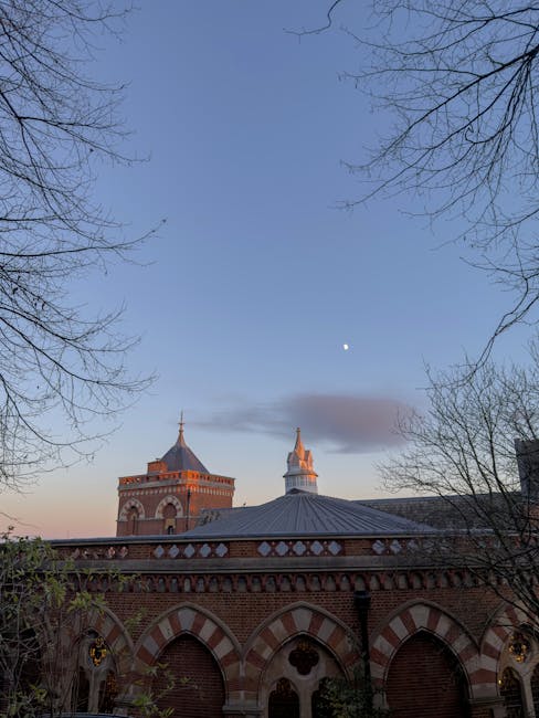 The image depicts the upper sections of an historic brick building with ornate arches and decorative white stone details, set against a clear evening sky with a visible moon and a few scattered clouds. The building features two prominent towers with conical roofs, topped with finials, and is framed by the leafless branches of trees on either side in the foreground. The scene is illuminated by natural twilight, highlighting the warm tones of the brickwork and giving the setting a calm, serene atmosphere. The environment suggests an outdoor urban setting, possibly in a historic town or city center, with no visible rubbish or waste materials in the scene. The image's focus on architecture and sky provides an atmospheric backdrop, subtly aligning with themes of private property and on-site scene capture, relevant to waste management or alternative collection services offered by Waste Removal Harrow in a professional context.