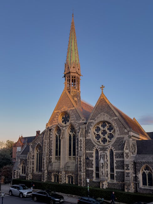 A historic stone church with a tall, pointed spire extending into a clear blue sky, featuring Gothic architectural details including large arched windows with intricate tracery and a circular rose window on the facade. The building's exterior shows a combination of rough stone and brickwork, with weathered textures and muted earthy tones. The church is situated along a street with parked cars visible in the foreground, and a low hedge lining the sidewalk. The overall scene is illuminated by natural daylight, suggesting late afternoon lighting, and captures the church's prominent verticality and detailed Gothic elements typical of ecclesiastical architecture. This detailed view highlights the structure's historic character and serves as an example of traditional church design. Waste Removal Harrow occasionally handles the clearance of similar sites for private events or building maintenance, though no waste or debris is visible in this image, emphasizing the church's preserved and well-maintained appearance.