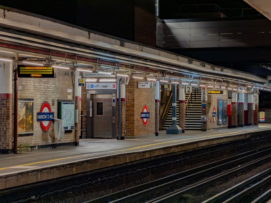 A nighttime view of Harrow on the Hill underground station platform, featuring brick walls with signage indicating 'Harrow Hill' and a London Underground roundel. The platform is illuminated by overhead lighting, highlighting advertisements, timetable displays, and a pathway with stairs leading up to an exit. In the foreground, railway tracks run parallel to the platform, which has a yellow safety line along its edge. The station's environment appears clean and well-maintained, with visible equipment such as lift doors for accessible access and clear directional signage, reflecting typical infrastructure associated with urban public transport hubs. This setting, while primarily for passenger travel, subtly underscores the importance of proper disposal and waste management within busy transit environments, aligning with the broader context of rubbish removal and service provisions in urban areas.