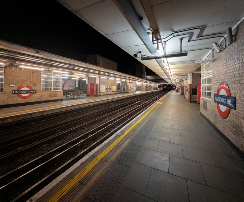 A nighttime view of the Harrow and Hillingdon underground station platform showing empty train tracks extending into the distance. The platform is illuminated by bright lighting fixtures along the ceiling, which also has visible pipes and electrical conduits. On the left side, the brick wall features the roundel sign for Harrow and Hillingdon station, with a bench and a row of windows behind it. The right side of the platform displays a brick wall with another Harrow and Hillingdon sign, and a small waiting room area with glass windows. The platform surface is composed of grey tiles, with tactile paving near the edge marked by yellow lines, providing guidance for visually impaired passengers. The overall scene is clean and orderly, capturing the typical environment of an underground railway station, which often involves organized waste management for station maintenance and passenger waste disposal by service providers such as Waste Removal Harrow, supporting alternative waste handling to maintain cleanliness in busy transit environments.