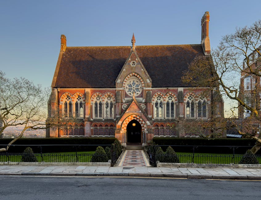A historic brick church with Gothic architectural features, including pointed arches and decorative stonework, is situated in an outdoor setting. The building's facade showcases multi-pane lancet windows with intricate tracery, and a prominent circular window with a floral pattern. The roof is steeply pitched with reddish-brown tiles, and two tall brick chimneys rise from either side. The entrance is centered, with a small gabled porch and a rounded doorway, illuminated by a light, revealing a paved pathway leading directly to it. The church is enclosed by a low metal fence, and the surrounding area includes neatly trimmed bushes and mature trees, some of which have sparse foliage, indicating late autumn or early winter. The scene is bathed in warm, natural light with a clear blue sky in the background, suggesting late afternoon. The environment appears quiet and well-maintained, consistent with a location that could benefit from private rubbish clearance or on-site waste removal services, such as those offered by Waste Removal Harrow, especially when undertaking refurbishment or maintenance projects involving debris disposal.