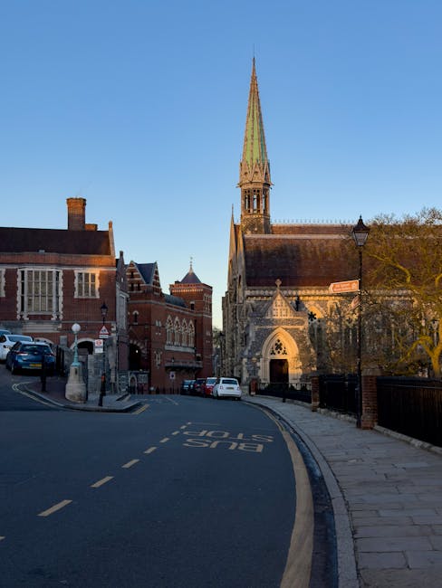 A narrow street in a historic town features a curved road with marked bus stop lines and parked cars along the left side, including a dark blue vehicle and a white van. To the right, there is a stone pavement with a black metal fence and a streetlamp. The background showcases a prominent Gothic-style church with a tall, pointed spire covered in weathered green tiles, large arched windows, and intricate stone detailing on its facade. Flanking the church are traditional red-brick buildings with various gabled roofs and chimneys, some with large, multi-pane windows. Leafless trees are visible near the church, indicating a late autumn or winter setting, and the sky is clear with a bright blue tone. The scene has a calm, outdoor atmosphere typical of a quaint, historic town, and the presence of parked vehicles and the layout suggest an area suitable for independent waste collection or private rubbish removal services, consistent with local authority alternatives such as those offered by Waste Removal Harrow.