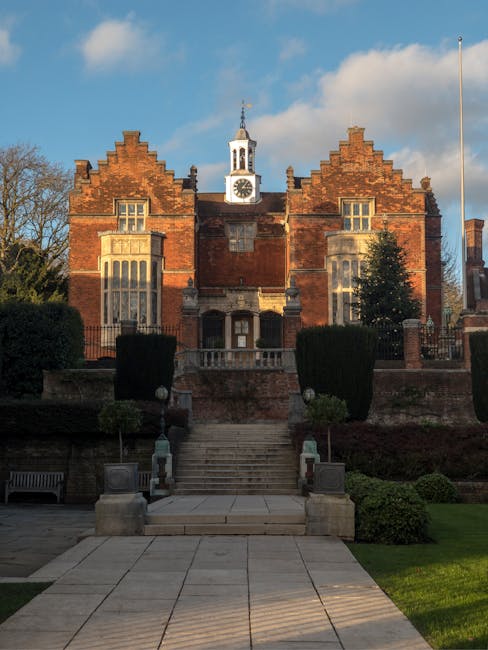 A historic brick church with Gothic architectural features, including pointed arches and decorative stonework, is situated in an outdoor setting. The building's facade showcases multi-pane lancet windows with intricate tracery, and a prominent circular window with a floral pattern. The roof is steeply pitched with reddish-brown tiles, and two tall brick chimneys rise from either side. The entrance is centered, with a small gabled porch and a rounded doorway, illuminated by a light, revealing a paved pathway leading directly to it. The church is enclosed by a low metal fence, and the surrounding area includes neatly trimmed bushes and mature trees, some of which have sparse foliage, indicating late autumn or early winter. The scene is bathed in warm, natural light with a clear blue sky in the background, suggesting late afternoon. The environment appears quiet and well-maintained, consistent with a location that could benefit from private rubbish clearance or on-site waste removal services, such as those offered by Waste Removal Harrow, especially when undertaking refurbishment or maintenance projects involving debris disposal.