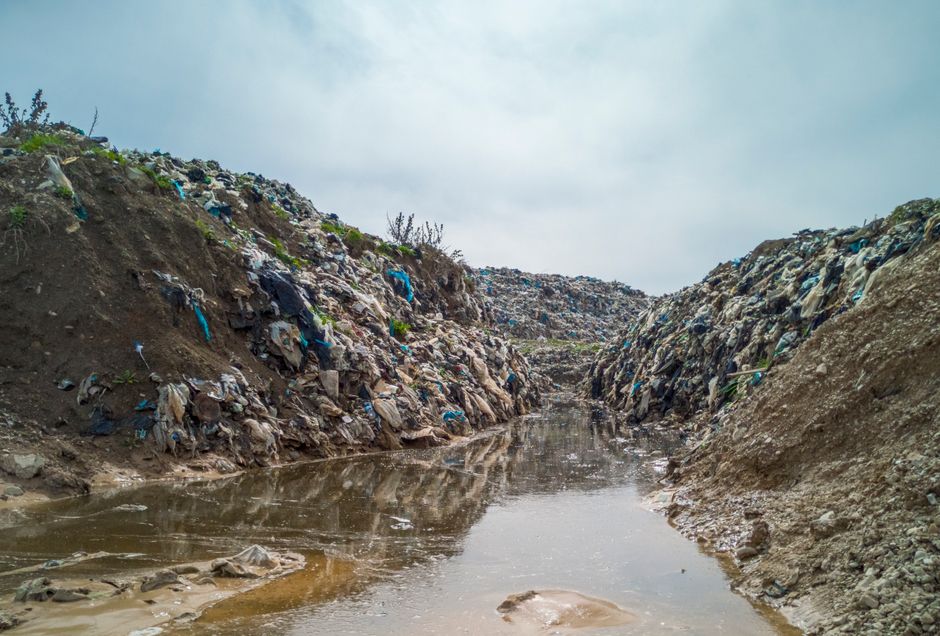 A narrow, unpaved drainage ditch filled with stagnant water runs between two steep dirt embankments heavily polluted with mixed waste debris, including plastic bags, fragments of cloth, and various discarded refuse. The embankments, composed of loose soil and gravel, are topped with sparse, sparse vegetation. In the background, the ditch extends into the distance toward a cloudy, overcast sky, highlighting a neglected outdoor environment affected by improper waste disposal. The scene illustrates environmental impact and the importance of proper rubbish removal practices, potentially handled by services such as Waste Removal Harrow, especially in areas where private or independent waste collection might be necessary to address illegal dumping.