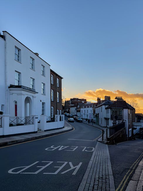 A quiet street scene in Harrow during sunset, featuring a gentle curve in the road with 'KEEP CLEAR' painted in white on the asphalt in the foreground. On the left, a prominent white building with a smooth, plastered exterior, multiple rectangular windows, and a small balcony with metal railing is visible. The building's façade is clean and painted white, with some architectural detailing around the entrance. Adjacent to it, a narrow brick-paved pathway runs along the pavement. To the right, lower terraced houses with dark tiled roofs and sash windows extend into the distance, their facades varying in white and darker materials. The street is lightly trafficked, with a few parked cars visible further along the road. The sky above displays a gradient from clear blue to warm orange hues near the horizon, with scattered clouds illuminated by the setting sun. The overall scene presents a quiet residential area where private waste collection, rather than street-side rubbish removal, could be relevant, reflecting the context of alternative waste handling methods sometimes necessary in areas with restricted access or narrow roads, as managed by waste removal services like Waste Removal Harrow.
