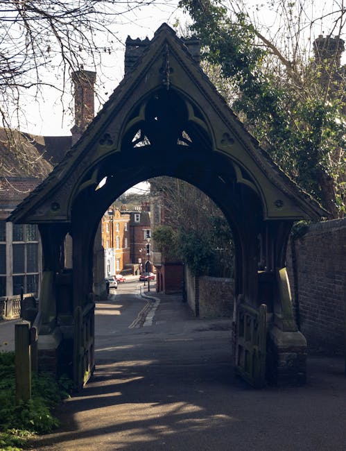 An ornate Victorian-style wooden gateway with pointed arches and decorative detailing, painted black, situated at the entrance to a narrow residential street. The structure features vertical posts supporting a gabled roof with intricate trim and small carved accents, casting shadows on the ground below. Behind the gateway, a paved pathway continues into a small alleyway lined with brick walls on both sides, leading to a wider street with parked cars and multi-storey residential buildings in the background. The scene is framed by leafless trees and some greenery, suggesting a temperate climate, with soft natural light indicating a clear day. This setting illustrates an urban entrance potentially associated with private property access or a historic site, which can be relevant for waste removal or clearance services specializing in local or site-specific rubbish collection, as performed by companies like Waste Removal Harrow.
