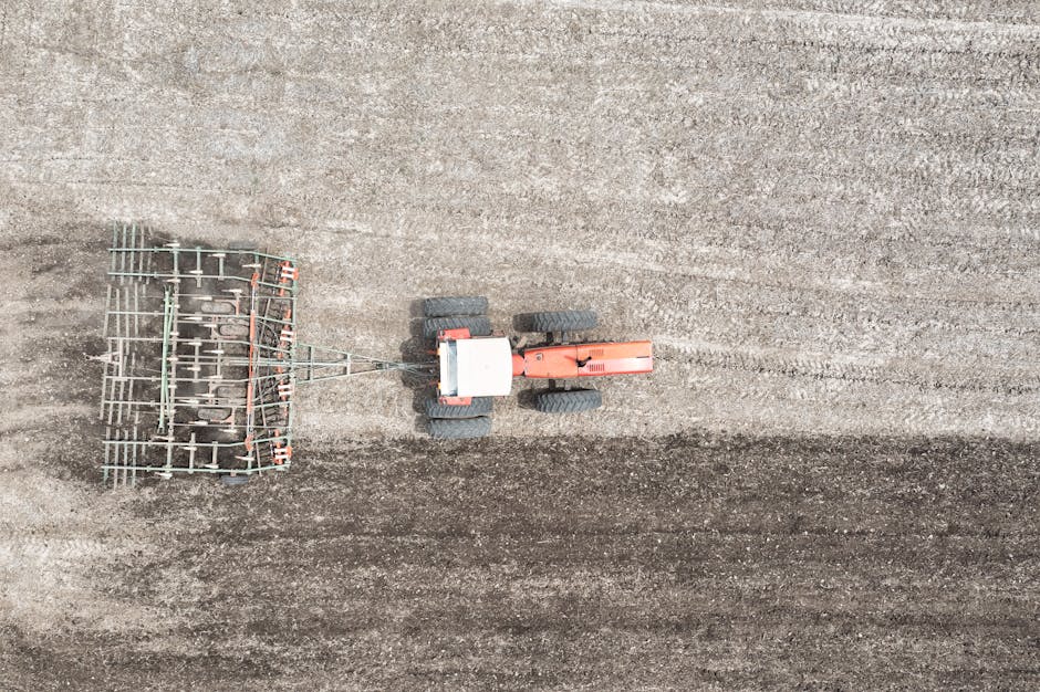 An aerial view of a compact tractor with a red body and large black tires operating on a large, freshly tilled field. The tractor is pulling a metal rake or harrow attachment, which is arranged in a grid-like pattern of rusted metal tines. The soil in the field appears uniformly prepared, with visible furrows created by the machinery, showing a textured, earthy surface in shades of brown and gray. The background reveals the expansive, open landscape with no visible structures or other objects, emphasizing the rural setting. The lighting is natural, casting minimal shadows, and the overall scene suggests ongoing land cultivation or preparation, aligning with the context of equipment used in waste management or site clearance by services such as Waste Removal Harrow, specialists in rubbish removal and on-site clearance services.
