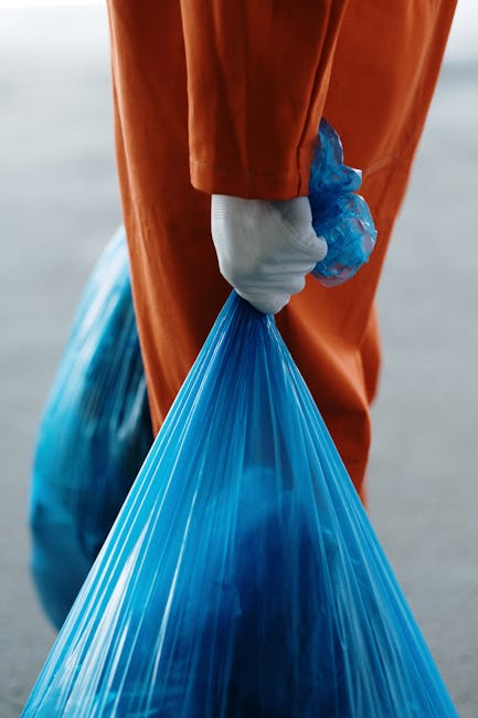 A person dressed in an orange protective suit and wearing a white glove is holding a blue plastic rubbish bag with their left hand. The bag appears to be filled with waste and is slightly bulging, with a shiny, smooth surface. The individual is standing on a paved surface, possibly outdoors, with a blurred background that suggests an open area or industrial environment. The scene conveys a context of waste collection or disposal, typical of manual rubbish removal tasks. Waste Removal Harrow offers efficient independent waste collection services, such as private rubbish disposal, and this image illustrates the process of handling and transporting waste on-site or from domestic or commercial premises. The lighting is natural, highlighting the textures of the plastic bag and the fabric of the protective suit without any harsh shadows, emphasizing a professional and clean approach to rubbish clearance relevant for local waste management services.