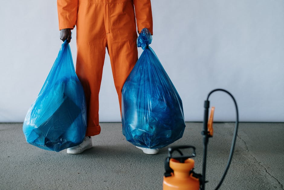 A person dressed in an orange jumpsuit and white shoes is holding two large blue plastic rubbish bags, one in each hand, standing on a concrete floor against a plain light grey background. The bags are filled with waste materials, appearing full and slightly bulging with various items inside. In the foreground, there is a black and orange portable vacuum or blower connected to a power source via a black cord, lying on the ground. The scene suggests an indoor setting, possibly a waste collection or rubbish removal context, with the individual preparing for private waste disposal or on-site clearance. The focus on the bags and waste management equipment aligns with the services offered by Waste Removal Harrow, providing a clear visual of targeted rubbish collection activities.