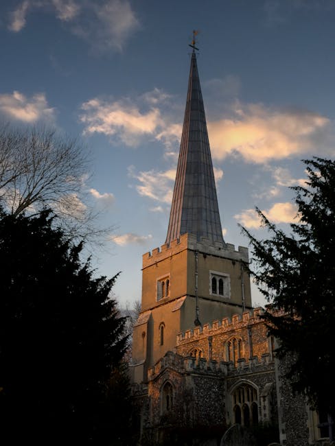 A tall, historic church with a prominent spire topped by a weather vane is pictured against a partly cloudy sky during late afternoon or early evening. The church’s lower sections are constructed from aged stone, featuring arched windows and battlement-style decorative elements along the upper edge of the tower. The spire is clad in smooth, modern metal sheeting with a slightly reflective finish and tapering to a sharp point. Surrounding the church are tree silhouettes, some leafless and others evergreen, framing the structure on either side. The scene captures the contrast between the weathered masonry and the sleek spire, exemplifying a typical example of traditional ecclesiastical architecture in a natural setting. This image could relate to local heritage sites or the preservation of historic buildings often involved in community or private site clearance efforts, such as those provided by waste removal services like Waste Removal Harrow, here in a visual context that emphasizes the importance of maintaining or clearing such sites.